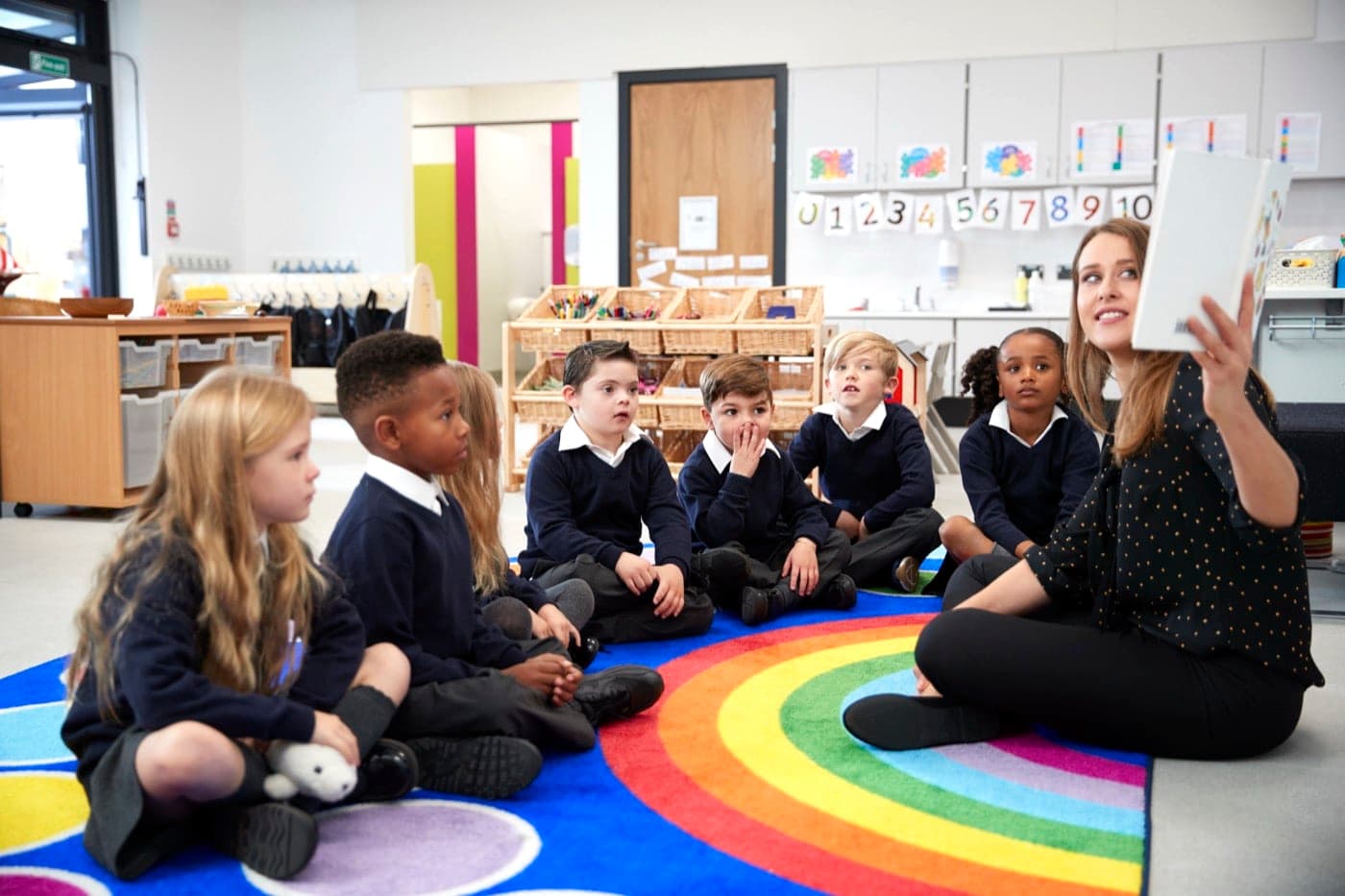 A teacher reading to a small group of young pupils sitting together on a colourful classroom rug